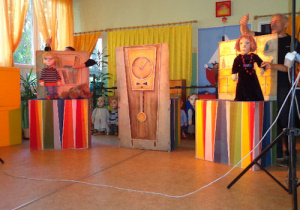 Po lewej stronie zdjęcia przed elementem scenografii przedstawiającym regał z książkami animator trzyma marionetkę przedstawiającą chłopca. Po prawej stronie przed elementem scenografii przedstawiającym regał z półkami animator trzyma marionetkę przedstawiającą kobietę w czarnej sukni z koralami na szyi.